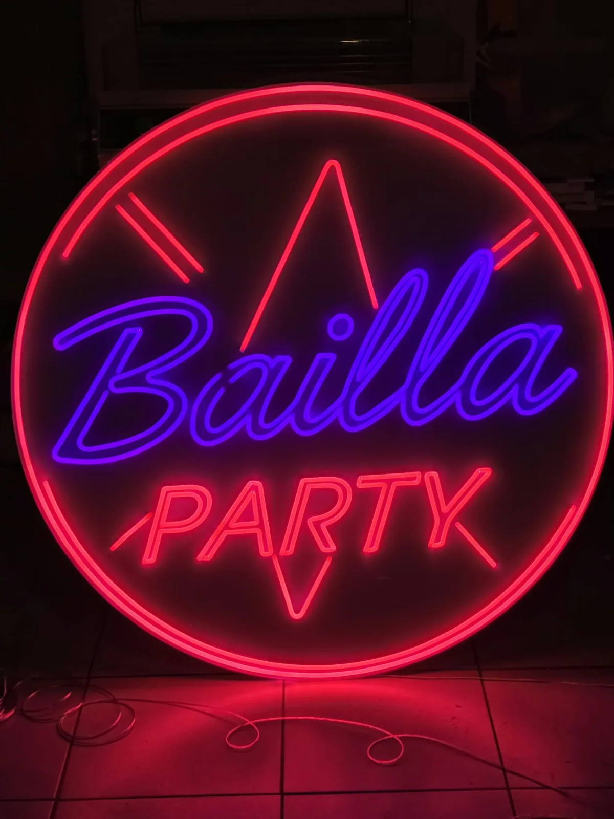 Okrągły neon LED „Bailla Party” — fioletowy napis w czerwonym pierścieniu z promieniami, świecący na ciemnej posadzce